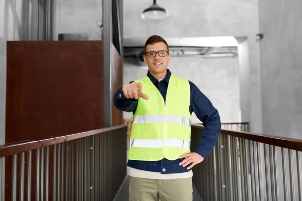 architecture, construction business and building concept - happy smiling male worker in safety west pointing to camera at office