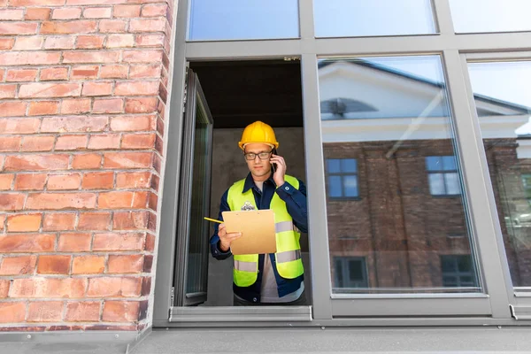 construction business and building concept - smiling male builder in helmet and safety west with clipboard calling on smartphone at window