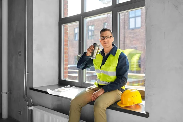 architecture, construction business and people concept - happy smiling male builder or architect with tumbler or thermo cup sitting on window sill at office