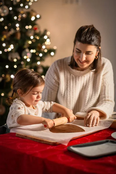 family, cooking and winter holidays concept - happy mother and baby daughter with rolling pin making gingerbread cookies from dough at home on christmas