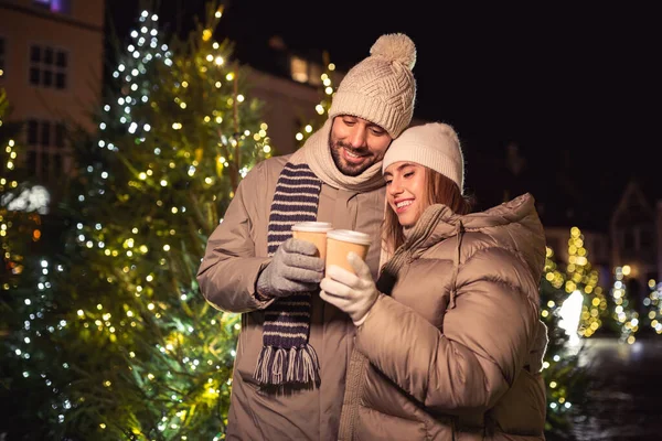 winter holidays, hot drinks and people concept - happy young couple with takeaway coffee cups over christmas tree lights in evening city