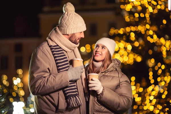 winter holidays, hot drinks and people concept - happy young couple with takeaway coffee cups in christmas city in evening