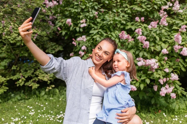 family, motherhood and people concept - happy mother and little daughter with smartphone taking selfie at summer park or garden