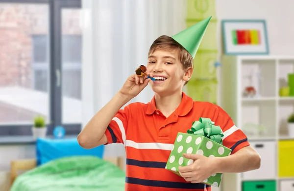 holiday, birthday and people concept - portrait of happy smiling boy in party hat with gift box and blower over home room background
