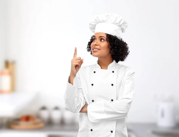 cooking, advertisement and food concept - happy smiling female chef in toque pointing finger up over restaurant kitchen background