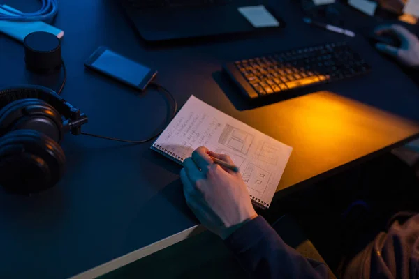 cybercrime, hacking and technology concept - close up of male hacker in dark room writing to notebook and using computer virus program for cyber attack
