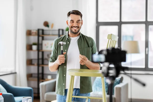 blogging, furniture restoration and home improvement concept - happy smiling man or blogger with camera, hammer and nail showing old wooden chair renovation and recording tutorial video