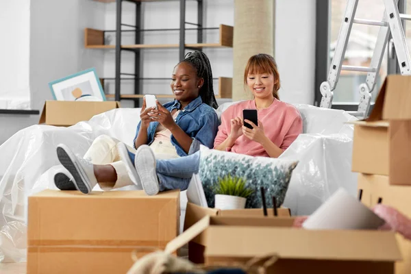 moving, people and real estate concept - happy smiling women with smartphones and boxes at new home
