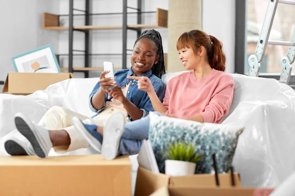moving, people and real estate concept - happy smiling women with smartphones and boxes at new home