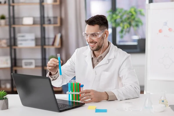 distance education, school and science concept - smiling male chemistry teacher in goggles and earphones with laptop computer having online class and showing test tube with chemical at home office
