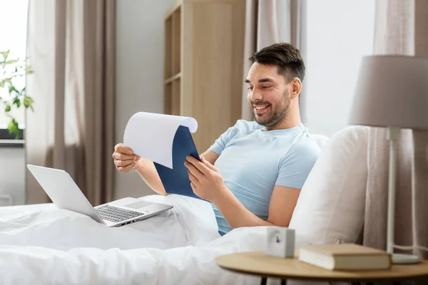 people, technology and remote job concept - man with laptop computer and clipboard working in bed at home bedroom