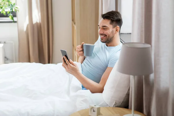 就寝时间和休息的概念 快乐的微笑的男人 带着智能手机在床上喝咖啡 — 图库照片