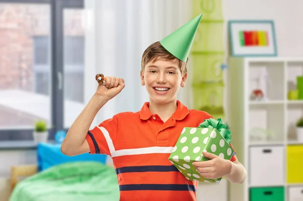 holiday, birthday and people concept - portrait of happy smiling boy in party hat with gift box and blower over home room background