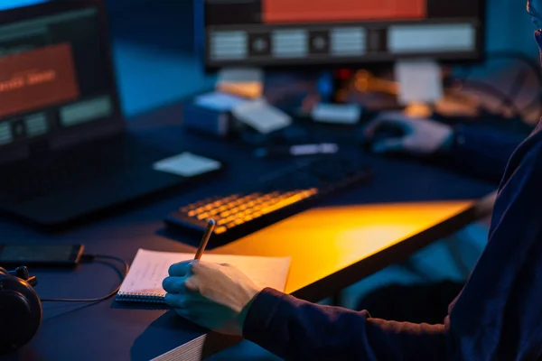 cybercrime, hacking and technology concept - close up of male hacker in dark room writing to notebook and using computer virus program for cyber attack