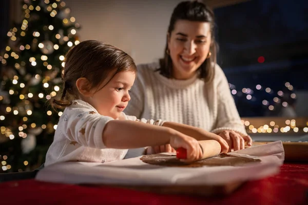 family, cooking and winter holidays concept - happy mother and baby daughter with rolling pin making gingerbread cookies from dough at home on christmas