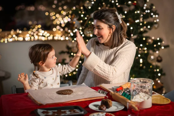 family, cooking and winter holidays concept - happy mother and baby daughter with rolling pin and dough for gingerbread cookies making high five gesture at home on christmas
