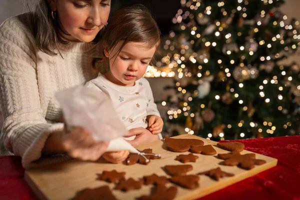 family, cooking and winter holidays concept - happy mother and baby daughter with icing in baking bag decorating gingerbread cookies at home on christmas