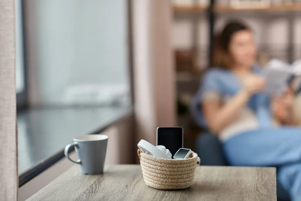 digital detox and leisure concept - close up of gadgets in basket on table and woman reading book at home