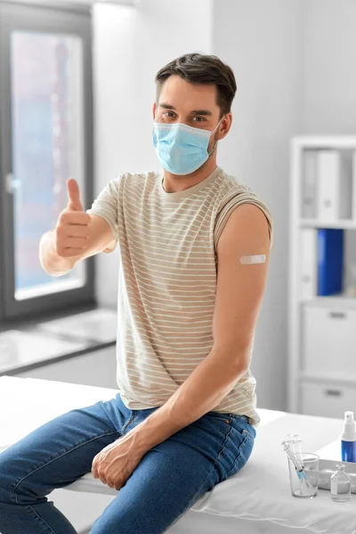 health, medicine and pandemic concept - happy male patient in mask with patch on his arm after vaccination showing thumbs up gesture at hospital
