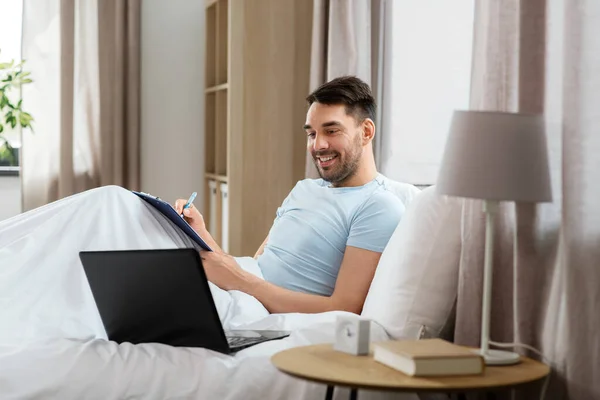 リモートジョブの概念 ラップトップコンピュータとクリップボードを持つ男は 自宅の寝室でベッドで働いています — ストック写真