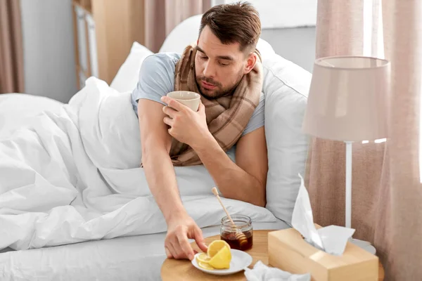 health, illness and people concept - sick man drinking hot tea with lemon and honey in bed at home