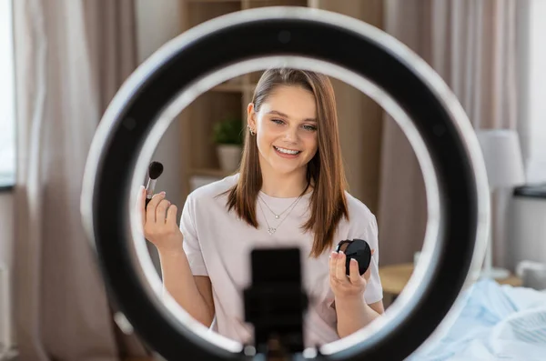 beauty blogging, technology and people concept - happy smiling girl blogger with ring light and smartphone applying make up at home
