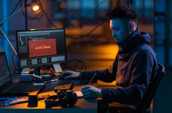 cybercrime, hacking and technology concept - male hacker in dark room writing to notebook and using computer virus program for cyber attack