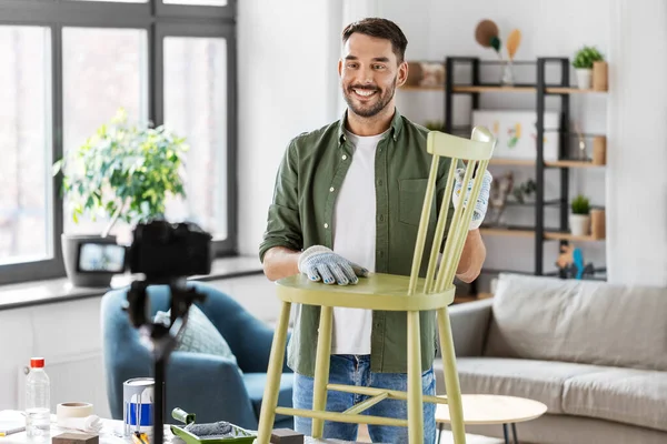 blogging, furniture restoration and home improvement concept - happy smiling man or blogger with camera showing old wooden chair renovation and recording tutorial video
