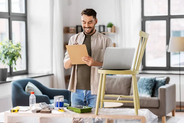 furniture restoration, diy and home improvement concept - happy smiling man with laptop and clipboard preparing old wooden chair for renovation