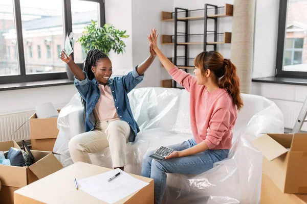 moving, finances and real estate concept - happy women with money, bills and calculator at new home making high five gesture