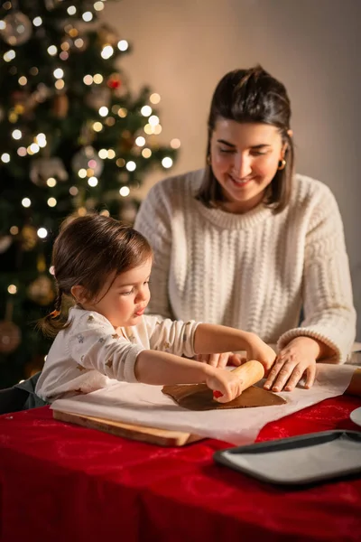 family, cooking and winter holidays concept - happy mother and baby daughter with rolling pin making gingerbread cookies from dough at home on christmas