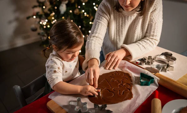 family, cooking and winter holidays concept - happy mother and baby daughter with mold making gingerbread cookies from dough at home on christmas