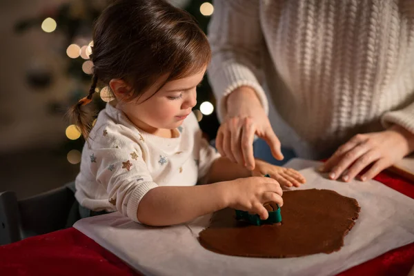 family, cooking and winter holidays concept - happy mother and baby daughter with mold making gingerbread cookies from dough at home on christmas