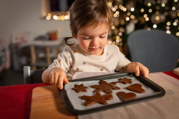 christmas, cooking and winter holidays concept - happy little baby girl with raw gingerbread cookies on baking tray at home