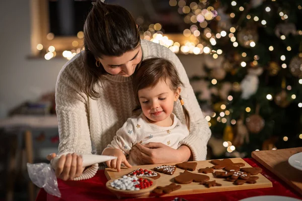 family, cooking and winter holidays concept - happy mother and baby daughter with icing in baking bag decorating gingerbread cookies at home on christmas