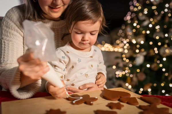 family, cooking and winter holidays concept - happy mother and baby daughter with icing in baking bag decorating gingerbread cookies at home on christmas