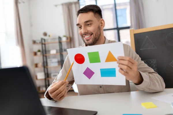 distance education, primary school and remote job concept - happy smiling male teacher with laptop and picture of geometric shapes in different colors having online class at home office