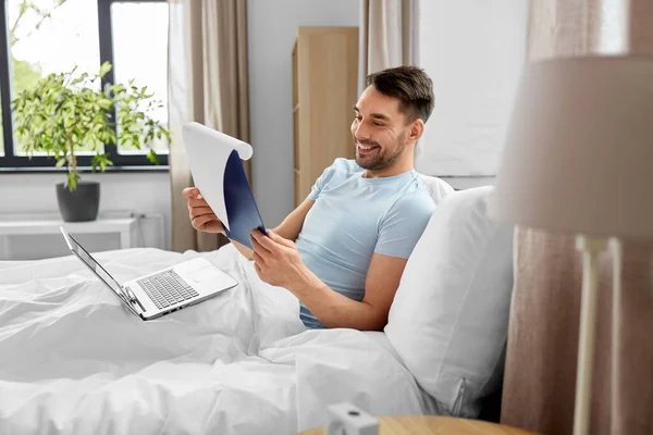 people, technology and remote job concept - smiling man with laptop computer and clipboard working in bed at home bedroom