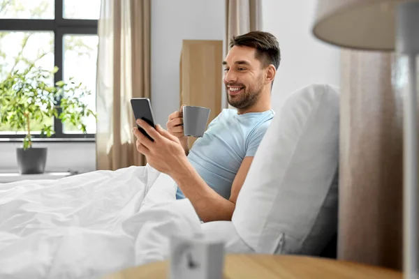 就寝時と休息の概念 自宅でベッドでスマートフォンのコーヒーを飲むと幸せな笑顔の男 — ストック写真