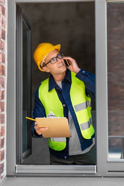 construction business and building concept - smiling male builder in helmet and safety west with clipboard calling on smartphone and looking out window
