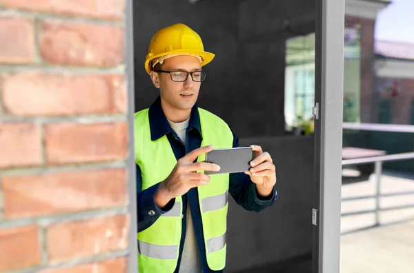 construction business and building concept - male builder in helmet and safety west with smartphone taking picture at window