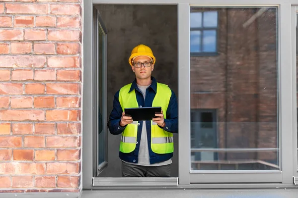 architecture, construction business and building concept - male architect in helmet and safety west with tablet pc computer in window