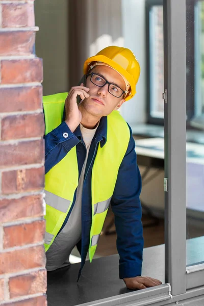 construction business and building concept - male builder in helmet and safety west calling on smartphone and looking out window