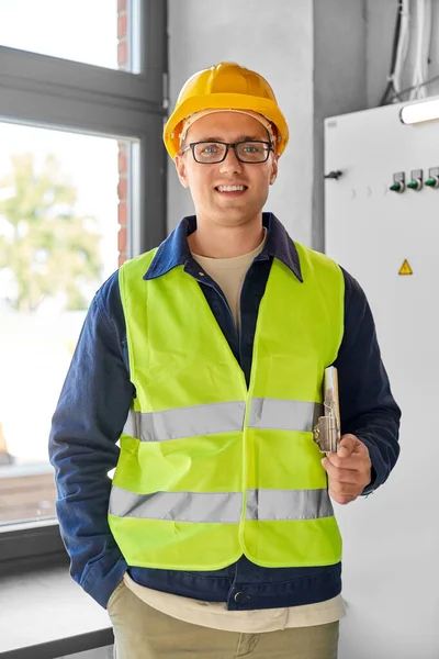 construction business and building concept - happy smiling male electrician or worker in helmet and safety west with papers on clipboard and pencil at electric board