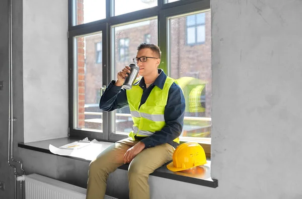 architecture, construction business and people concept - male builder or architect with tumbler or thermo cup sitting on window sill at office