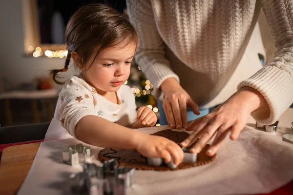 family, cooking and winter holidays concept - happy mother and baby daughter with mold making gingerbread cookies from dough at home on christmas