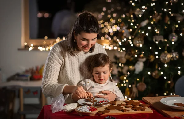 family, cooking and winter holidays concept - happy mother and baby daughter decorating gingerbread cookies at home on christmas