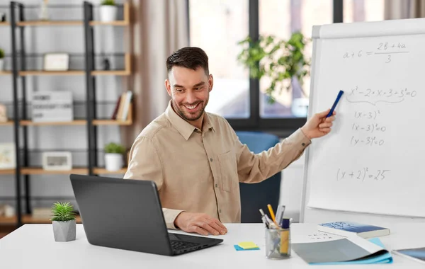 distant education, school and remote job concept - happy smiling male math teacher with laptop compute showing equation on flip chart having online class or video call at home office