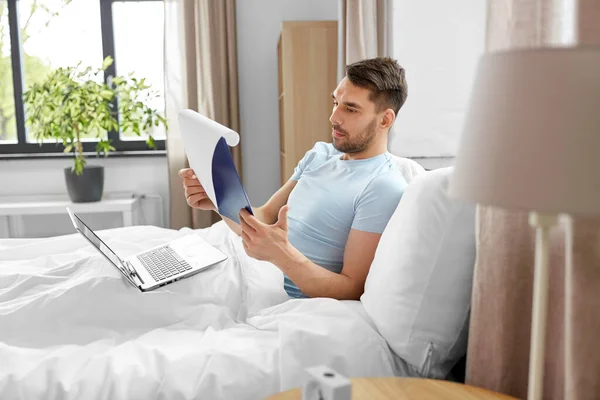 people, technology and remote job concept - man with laptop computer and clipboard working in bed at home bedroom