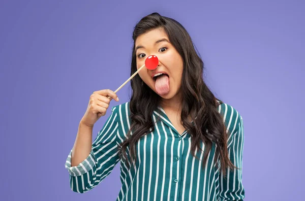 red nose day, party props and photo booth concept concept - funny woman with clown nose sticking out tongue over violet background